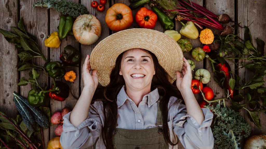 Gardener holding on to straw hat grinning wide with her autumn harvest all around her - Home for the Harvest - harvesting season in the vegetable garden