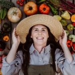 Gardener holding on to straw hat grinning wide with her autumn harvest all around her - Home for the Harvest - harvesting season in the vegetable garden