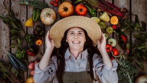 Gardener holding on to straw hat grinning wide with her autumn harvest all around her - Home for the Harvest - harvesting season in the vegetable garden