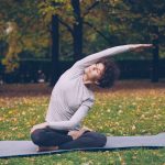 Slim young lady is doing sequence of yoga asanas sitting on mat in park alone on warm autumn day with beautiful grass and trees around her. Recreation and sport concept.